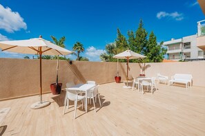 Eine Terrassengruppe im Freien mit weißem Mobiliar, zwei Sonnenschirmen und Pflanzkübeln.
