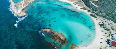 An aerial view of a coastal area with a beach, clear blue waters, and scattered buildings.