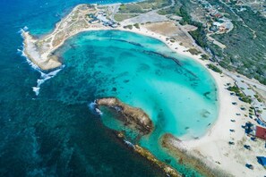Vista aérea de una zona costera con playa, aguas azules cristalinas y edificios dispersos.