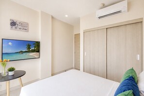 Ein Hotelzimmer mit einem Bett, einem Fernseher, der eine Strandansicht zeigt, einer Wand-Klimaanlage und einer Tür zu einem weiteren Raum.