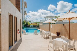 Ein Poolbereich mit Liegestühlen und Sonnenschirmen, einem Feuerlöscher und Blick auf ein Gebäude mit Pool im Hintergrund.