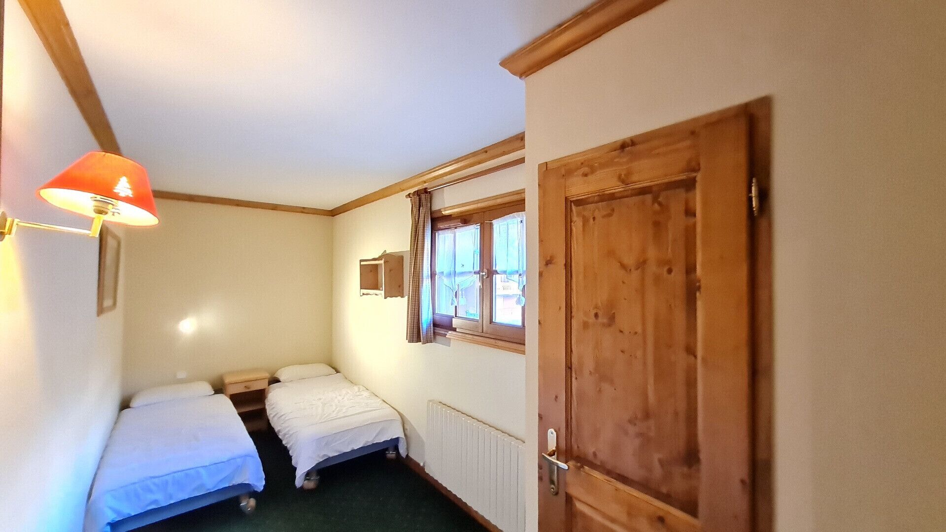 A small, cozy room with two single beds, a wooden door, a window with curtains, and a wall-mounted lamp.