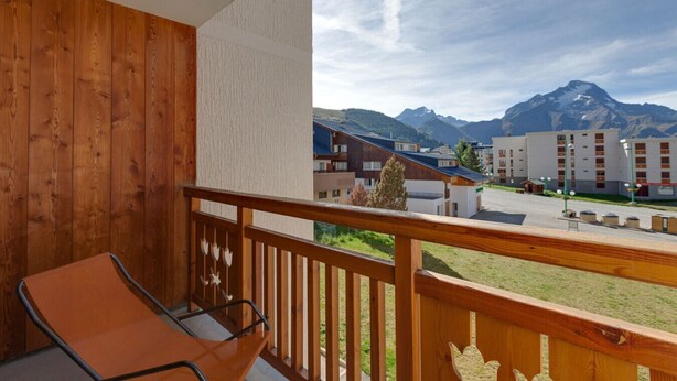 A balcony with a wooden railing, a single chair, and a view of a mountain range and buildings in the background.