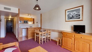 A compact kitchen with wooden cabinets, a dining area with a table and chairs, a television on a wooden stand, and a framed picture on the wall.