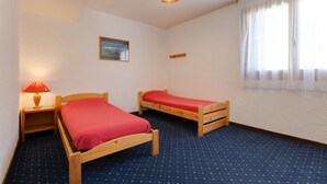 A room with two wooden beds, a single nightstand with a lamp, a framed picture on the wall, and a window with sheer curtains.