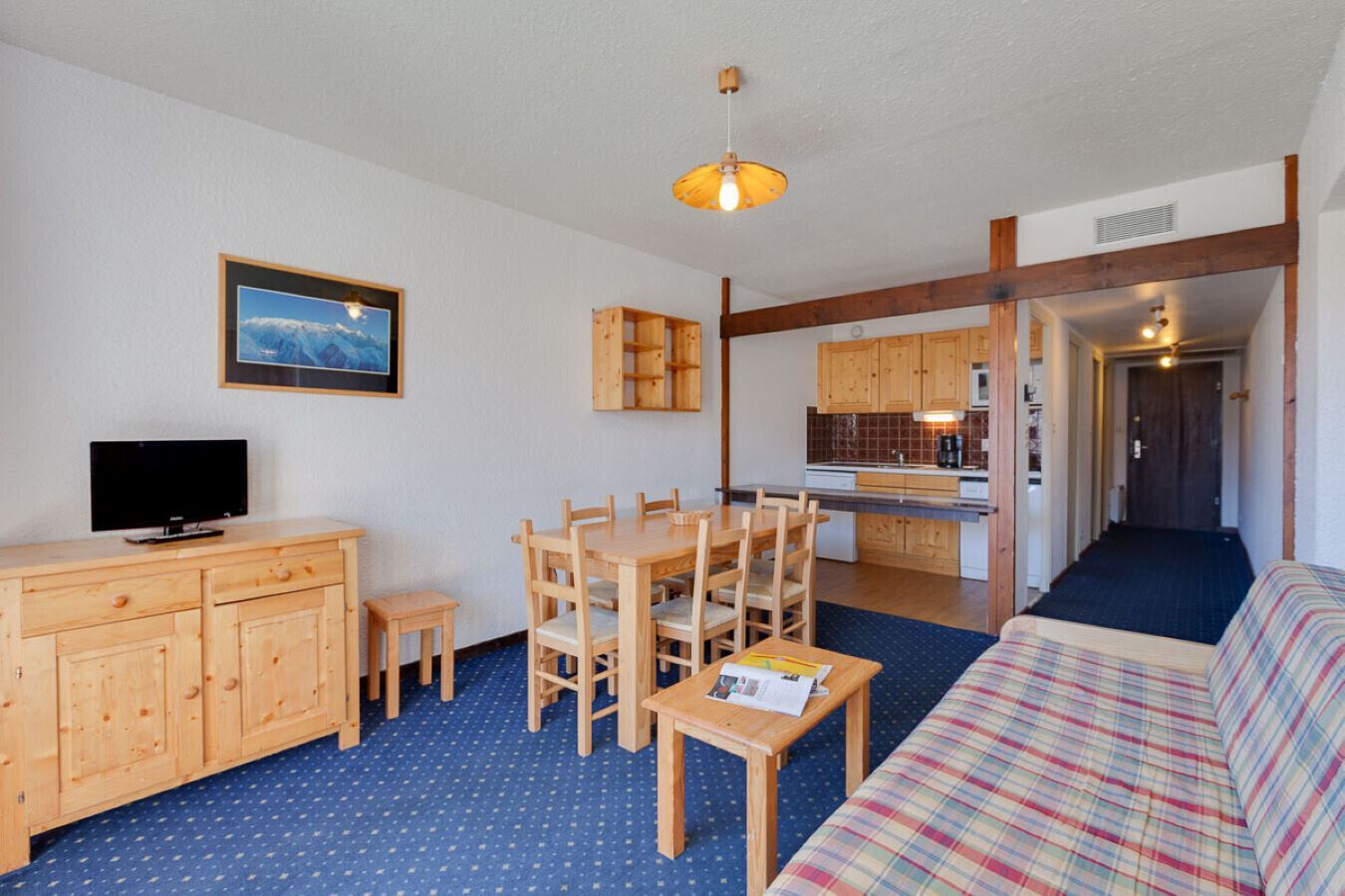 Ein Zimmer mit Esstisch, Stühlen, einer Couch, einem Fernseher, Holzschränken und einer Küchenzeile.