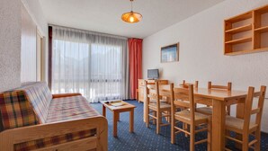 A room with a sofa, dining table, chairs, a small table, a television, and a window with curtains.