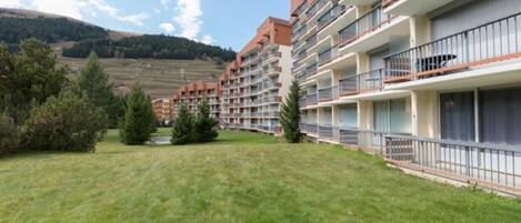 Un hotel de varios pisos con balcones, un césped y montañas al fondo.