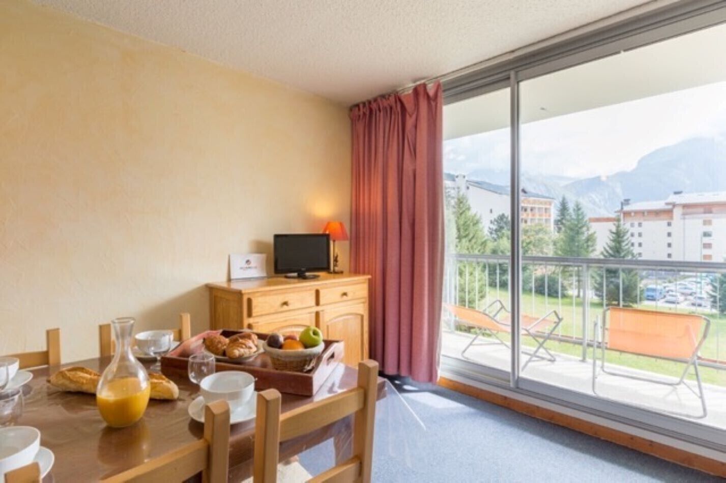 Ein Hotelzimmer mit Frühstückstisch, Fernseher auf einer Holzkommode und einem Balkon mit Blick auf Gebäude und Berge.