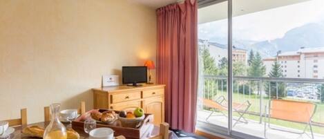 Ein Hotelzimmer mit Frühstückstisch, Fernseher auf einer Holzkommode und einem Balkon mit Blick auf Gebäude und Berge.