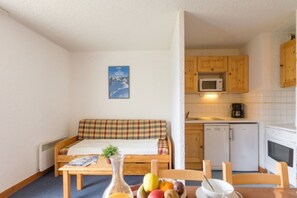 Um quarto acolhedor com um sofá, uma mesa de jantar com frutas, uma cozinha com armários de madeira e uma pequena planta.