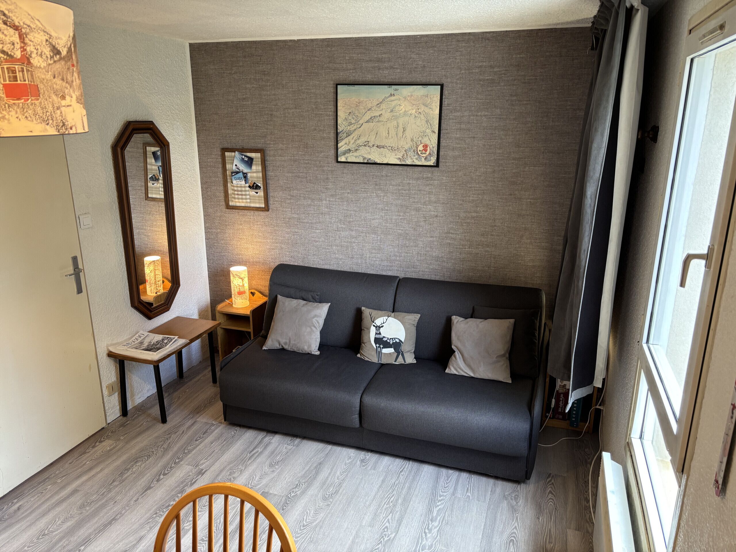 A cozy living room with a dark grey sofa, a wooden chair, a small table with a lamp, and a mirror.