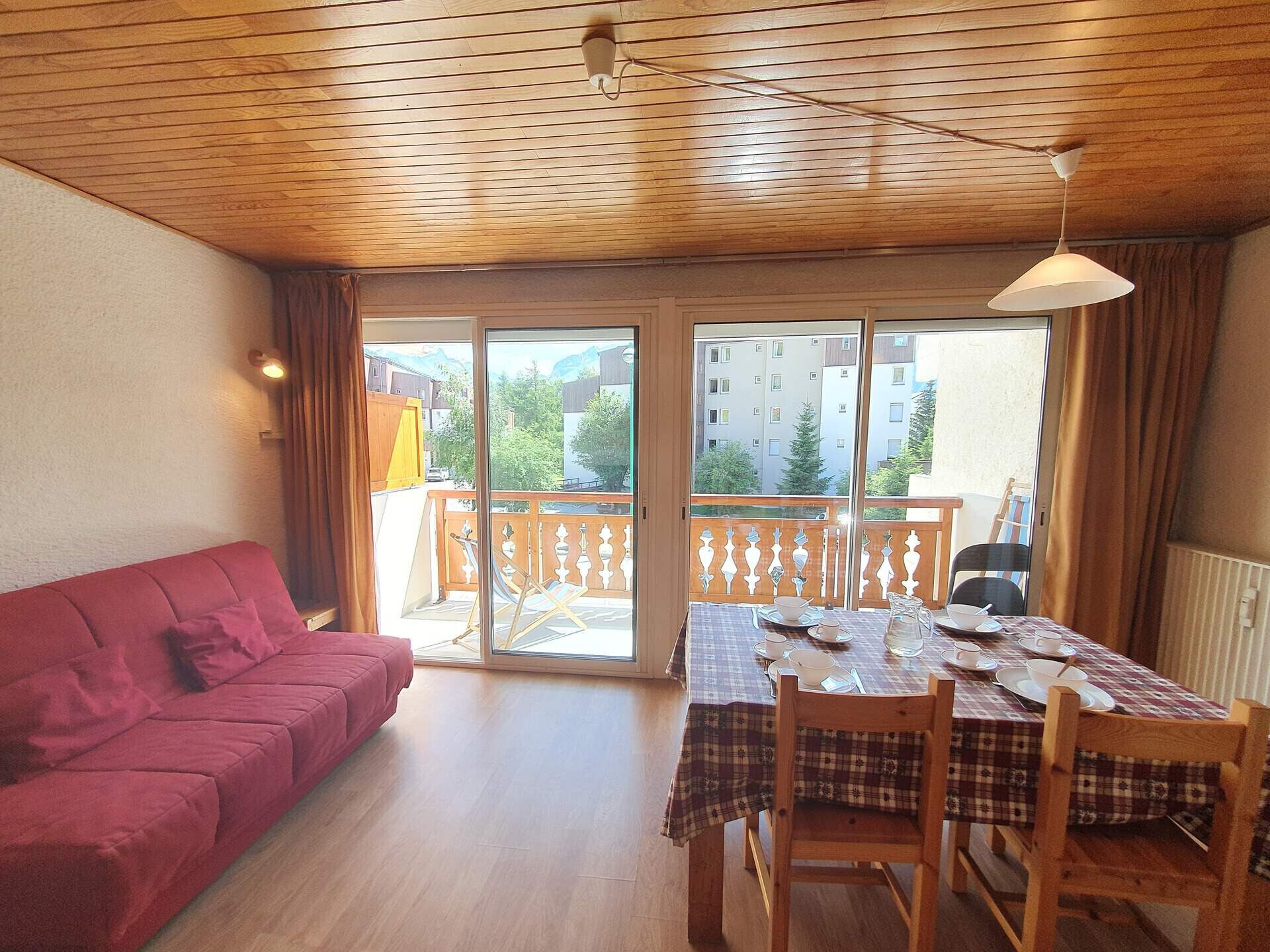 Una habitación con un sofá rojo, una mesa de comedor con platos y vasos, sillas de madera, un balcón con barandilla y vistas a edificios y árboles.