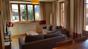Un salon comprenant un canapé, une table basse et un meuble pour la télévision. Il y a de grandes fenêtres avec des rideaux, un radiateur et une lampe.