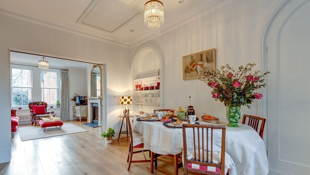 Et spiseområde med et hvidt bord dækket til fire personer, omgivet af træstole. En lysekrone hænger fra loftet, og der er en vase med blomster på bordet.