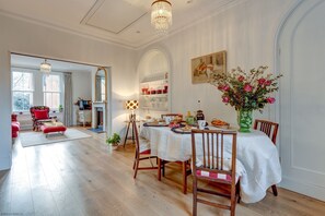 A dining area with a white table set for four, surrounded by wooden chairs. A chandelier hangs from the ceiling, and a vase with flowers is on the table. - University Parks View | Oxford Holiday Let | 1 Bed & Sleeps 2 (Oxford City Centre)