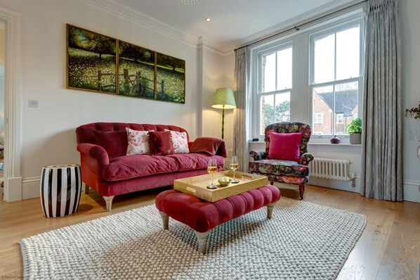A living room with a red sofa, a wooden coffee table, a patterned armchair, and a large window with curtains. - University Parks View | Oxford Holiday Let | 1 Bed & Sleeps 2 (Oxford City Centre)