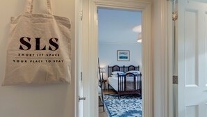 En døråbning ind til et soveværelse med en seng, et natbord og en vægmonteret lampe. En beige totebag med teksten "SLS" og "Short Let Space" hænger på væggen.