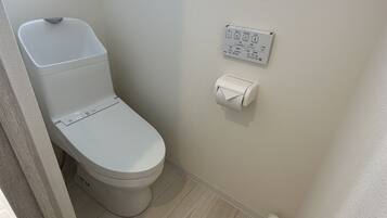 Inodoro blanco con botón de descarga y dispensador de papel higiénico en la pared.