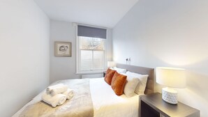A neatly arranged bedroom with a bed, pillows, a bedside table with a lamp, a window with blinds, and a framed picture on the wall. - Oxford Bolthole | Oxford Holiday Let | 2 Beds & Sleeps 4 (Oxford)