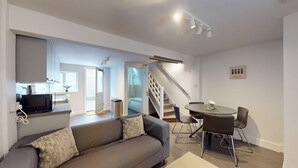 A modern living room with a grey sofa, a dining table with chairs, a staircase, and a kitchen area with a microwave. - Oxford Bolthole | Oxford Holiday Let | 2 Beds & Sleeps 4 (Oxford)