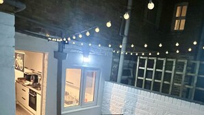 An outdoor patio with a round table and chairs, string lights, and a brick wall. - Oxford Bolthole | Oxford Holiday Let | 2 Beds & Sleeps 4 (Oxford)