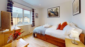 Ein Schlafzimmer mit einem hölzernen Bett, weißer Bettwäsche, einem Nachttisch mit Lampe, einem Fenster mit Vorhängen und Blick auf Häuser.