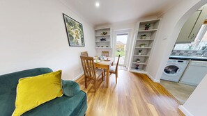 A compact living space with a dining area, a sofa with a yellow pillow, wooden flooring, a kitchen with a washing machine, and a view through an open door. - Cherry Tree Cottage | Bicester Holiday Let | 3 Beds & Sleeps 5 (Bicester)
