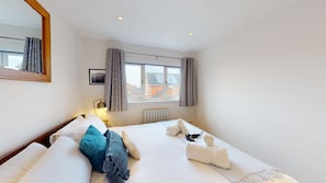 A hotel room with a bed, a nightstand, a lamp, a window with curtains, a radiator, and a mirror. - Cherry Tree Cottage | Bicester Holiday Let | 3 Beds & Sleeps 5 (Bicester)