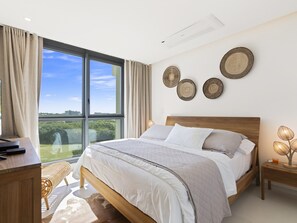 A modern bedroom with a large window, a wooden bed, a desk, and a bedside table with a lamp. - B-404 Stunning one bedroom corner apartment (Cupecoy)