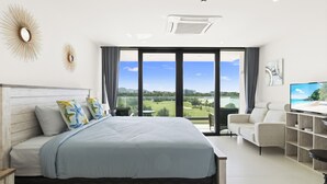 A modern hotel room with a large bed, a desk with a flat-screen TV, and a view of a golf course and beach.