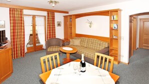 Ein gemütliches Zimmer mit einer Couch, einem kleinen Tisch mit Flasche und Gläsern, einem Sessel, einem Bücherregal und einem Fernseher.