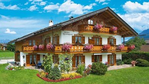 A two-story wooden building with balconies, a garden with flowers, and mountains in the background.