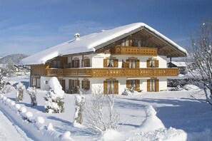 Un chalet in legno a due piani con un balcone, circondato da alberi innevati e un cielo sereno.