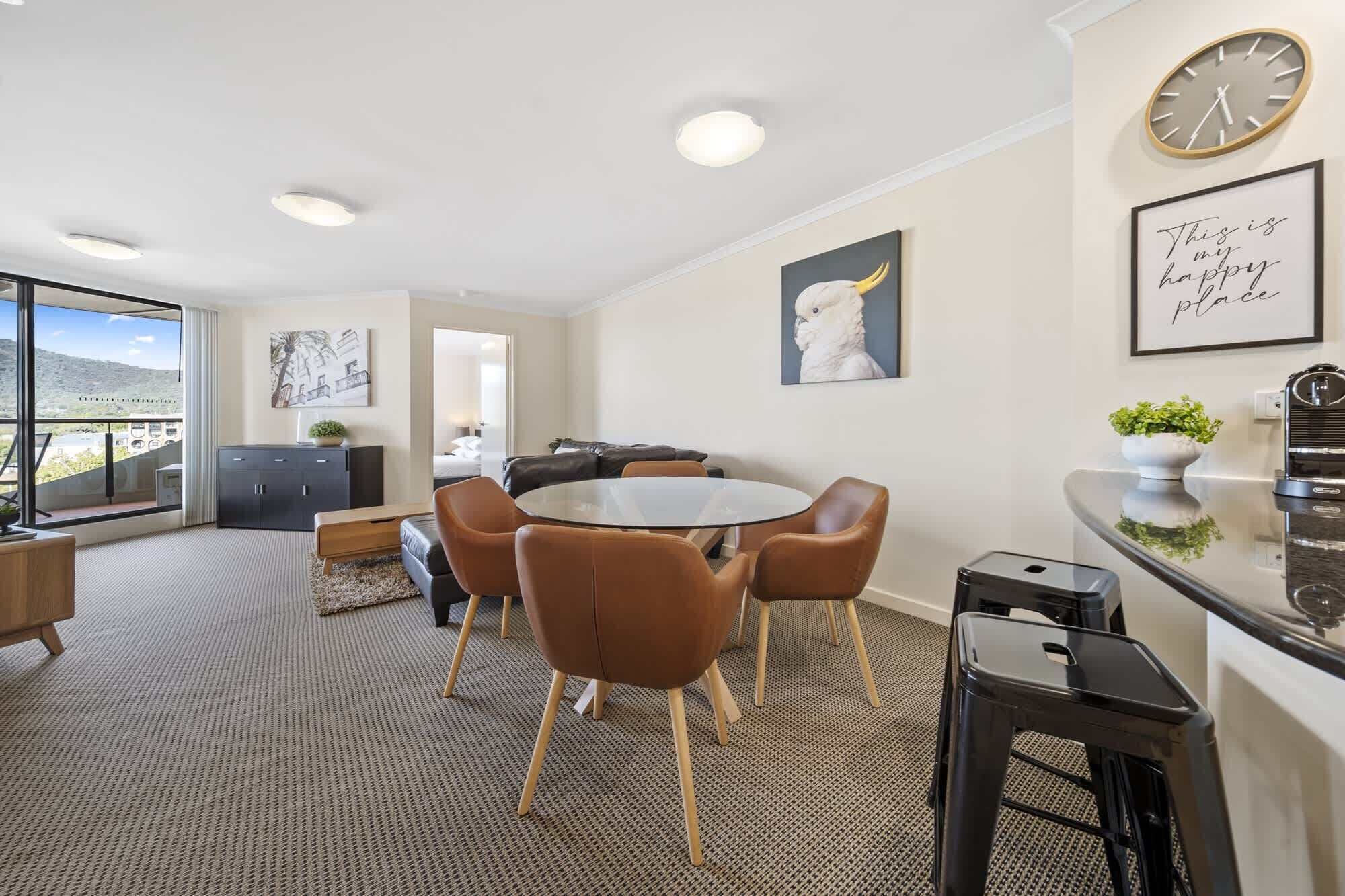 Een moderne eetruimte met een ronde tafel en zes stoelen, een keuken met een klok en kunstwerken in lijsten, en uitzicht door een groot raam.