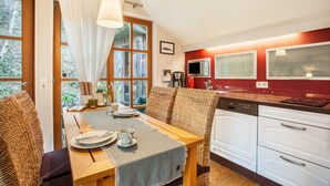 Un coin repas avec une table dressée pour deux personnes, une cuisine avec des appareils électroménagers blancs et une fenêtre avec des rideaux.
