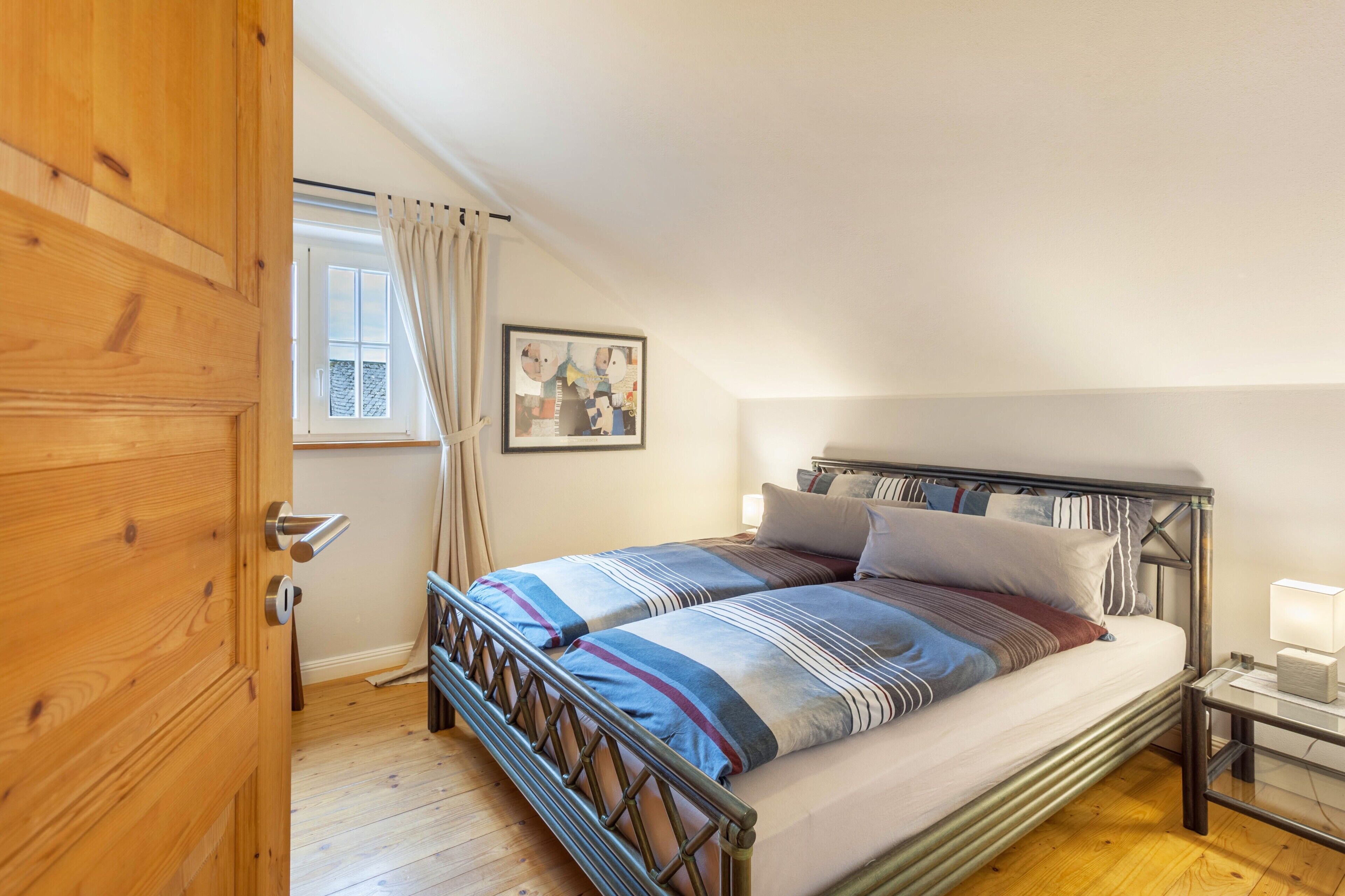A bedroom with a wooden door, a window with curtains, a bed with striped bedding, a bedside table with a lamp, and a wall-mounted picture.