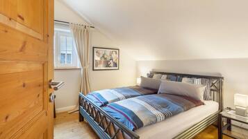 A bedroom with a wooden door, a window with curtains, a bed with striped bedding, a bedside table with a lamp, and a wall-mounted picture.