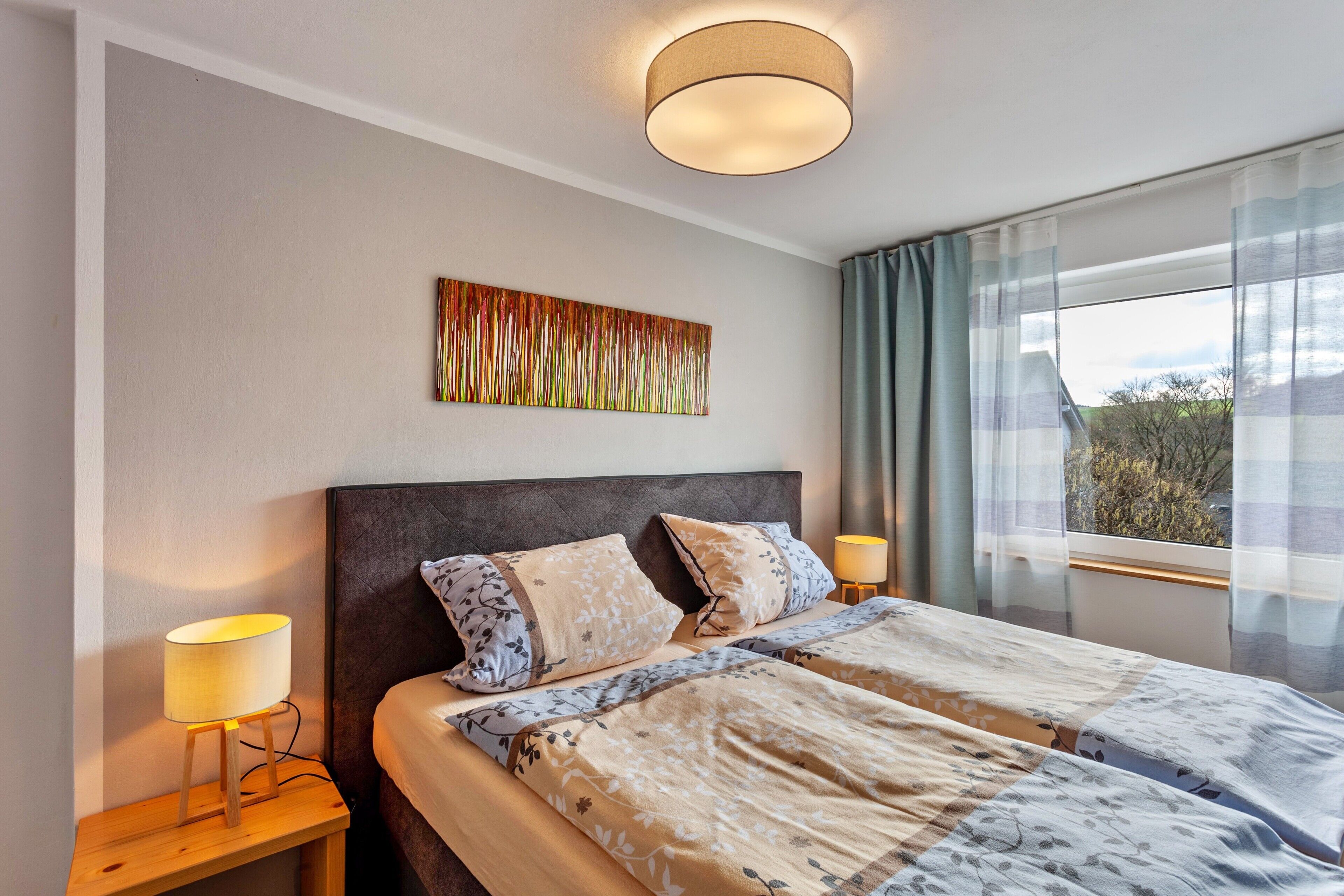 Ein Hotelzimmer mit einem Bett, zwei Nachttischlampen, einem Wandbild und einem Fenster mit Vorhängen.