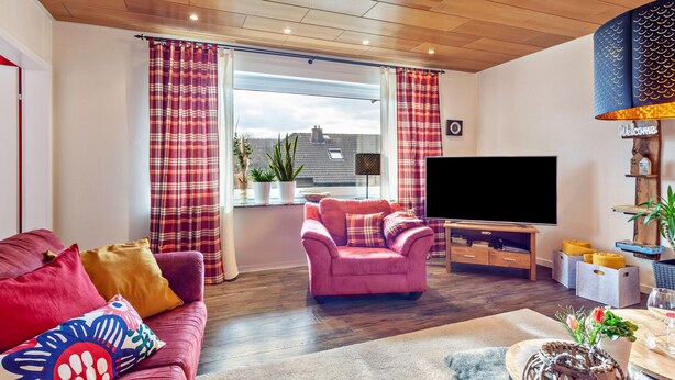 A living room with a sofa, armchair, TV, wooden ceiling, and a window with curtains.