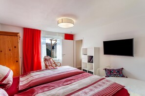 Un dormitori amb llit, cadira, armari, televisió i finestra amb cortines.