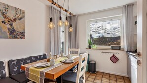 A dining area with a wooden table, white chairs, a bench, and a view of a tree through a window.