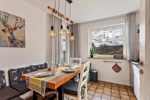 Un coin repas avec une table en bois, des chaises blanches, un banc et une vue sur un arbre par la fenêtre.
