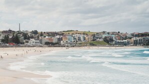 A beach with waves, a sandy shore, and a cityscape with buildings in the background. - Bondi Pacific Studio - Managed by X Hospitality (Bondi Beach)
