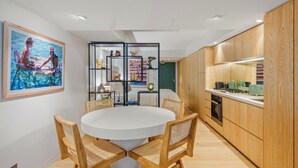 A modern kitchen with wooden cabinets, a dining area with a round table and chairs, and a wall-mounted shelf with decorative items. - Bondi Pacific Studio - Managed by X Hospitality (Bondi Beach)