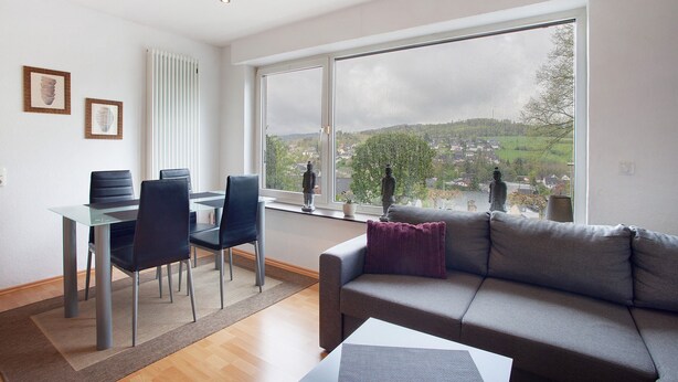 A modern living room with a sofa, a dining table with chairs, and a large window offering a view of a town. - Apartment "Keil" with Mountain View, Private Terrace & Wi-Fi (Balve)