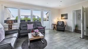 Un salon spacieux comprenant un canapé, un fauteuil et une table basse, de grandes fenêtres et une porte donnant sur une autre pièce.