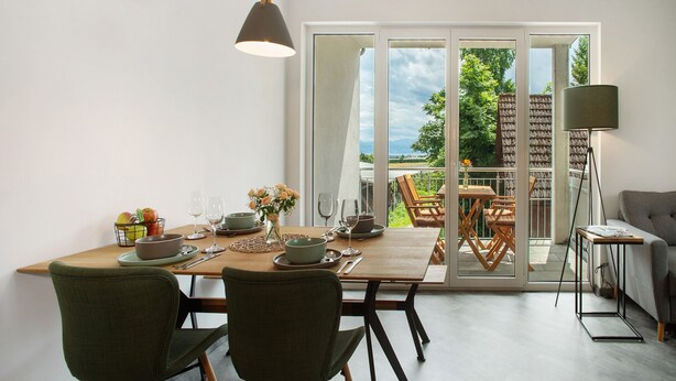 A dining area with a table set for four, chairs, a vase with flowers, and a view through a glass door to an outdoor seating area.