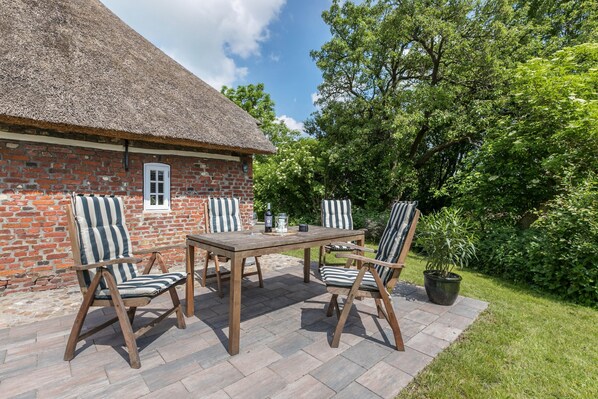 A brick building with a thatched roof, a wooden table, and striped chairs in a garden setting. - Holiday Home "Luis - Hus" with 2 Private Terraces, Garden, and Wi-Fi (Dagebüll)