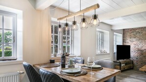 A dining area with a wooden table, black upholstered chairs, hanging light fixtures, and a brick wall. - Holiday Home "Luis - Hus" with 2 Private Terraces, Garden, and Wi-Fi (Dagebüll)